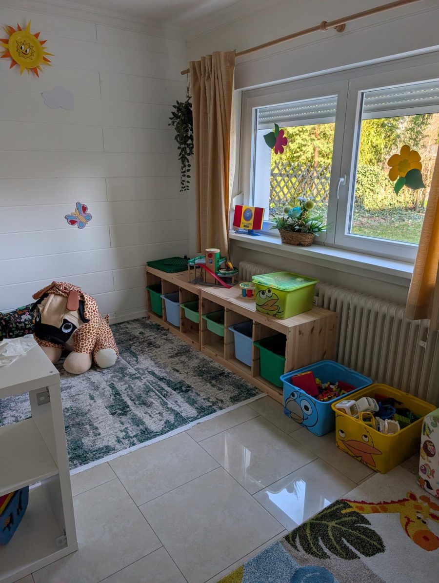 Niedriges Holzregal mit bunten Spielkisten vor dem Fenster, dahinter der Garten