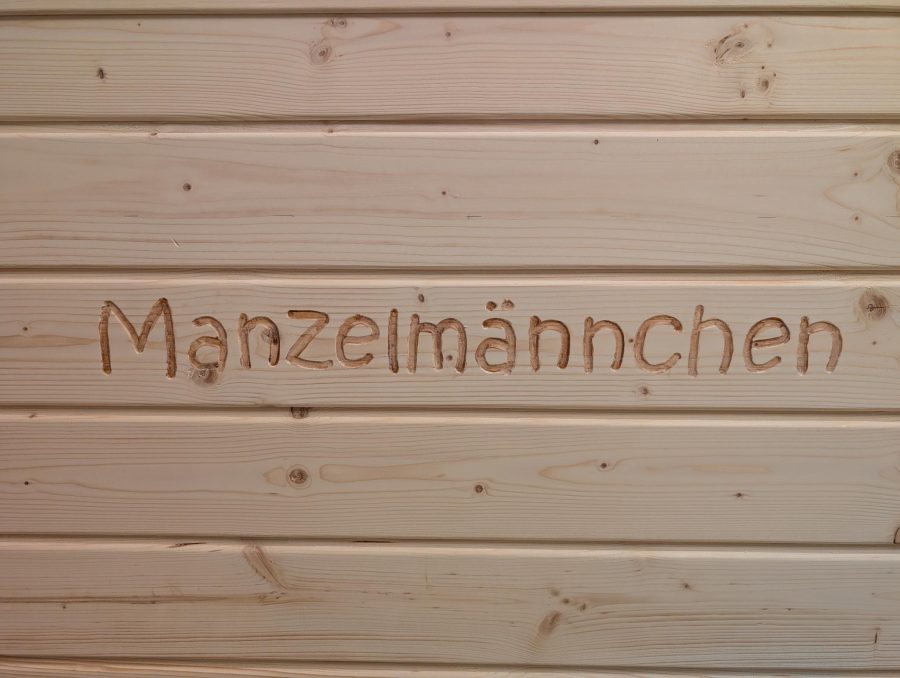 In eine helle Holzwand gefräster Schriftzug „Manzelmännchen“